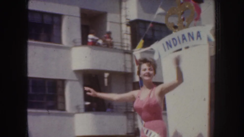 1952 California 8mm frame showing a smiling sash-wearing woman gesturing beside an Indiana sign and pennants.