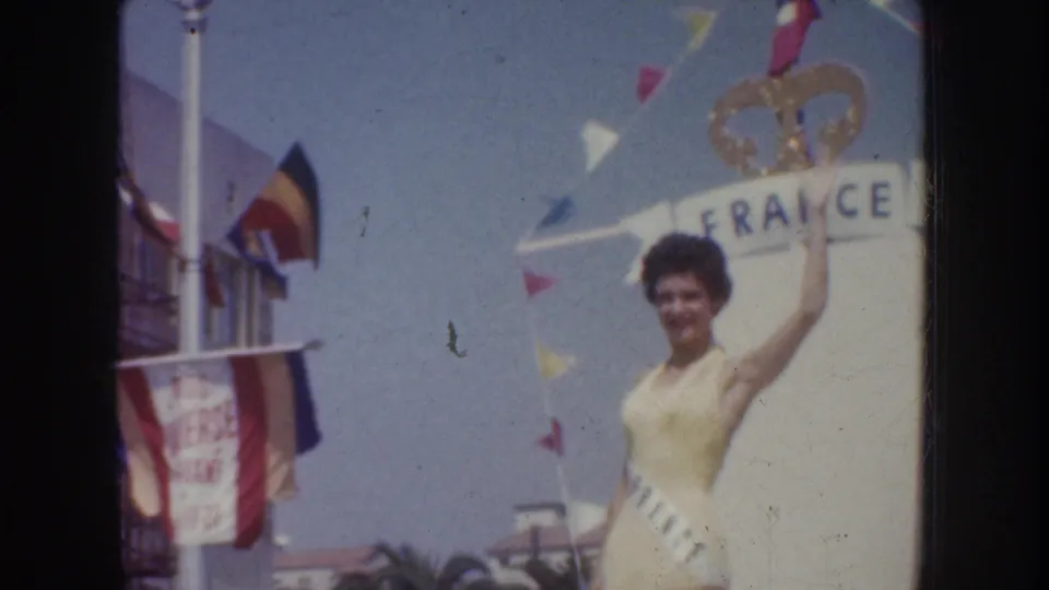 1952 California parade frame with a sash-wearing woman waving beside a France sign, pennants, and nearby flags.