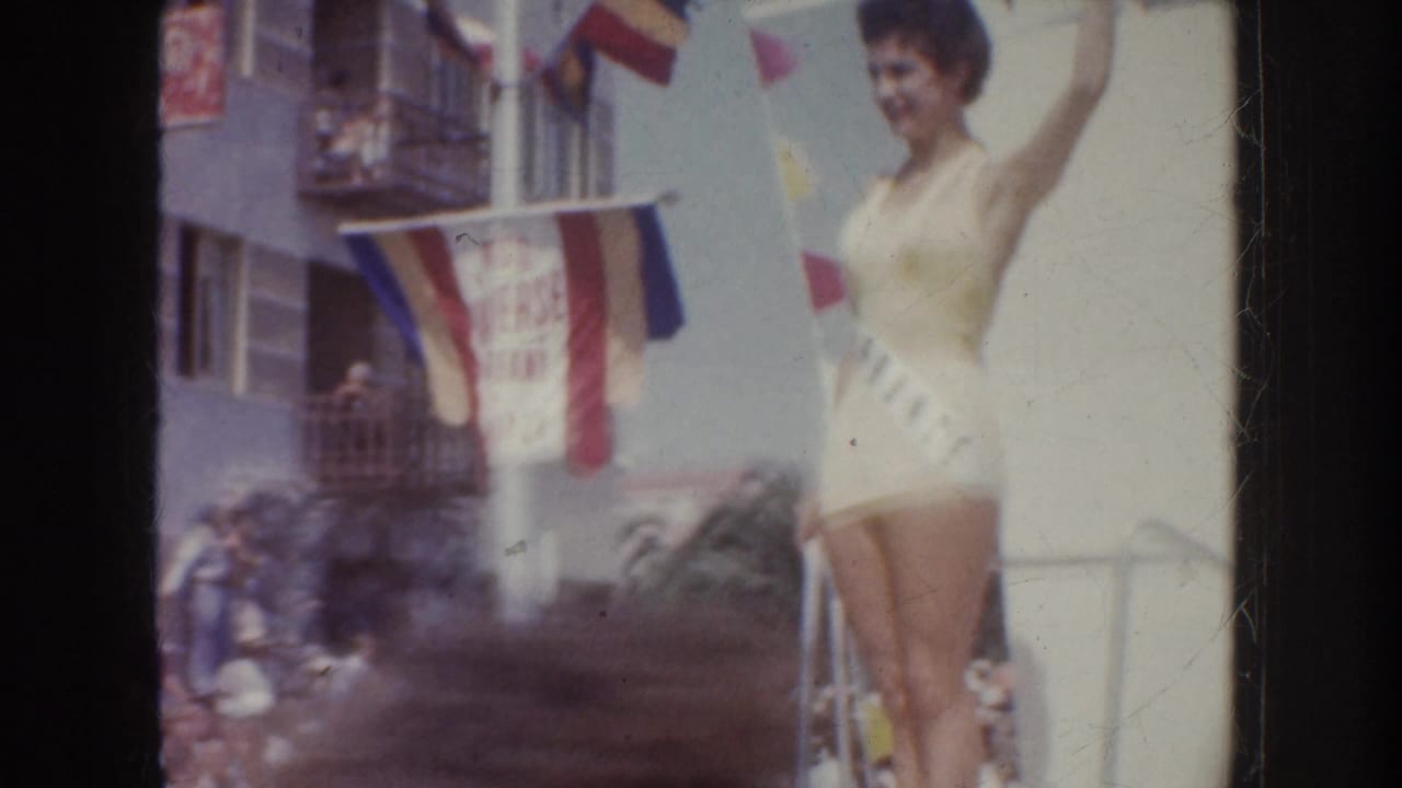 1952 California home-movie frame showing a swimsuit contestant waving near parade flags, with spectators and buildings behind her.