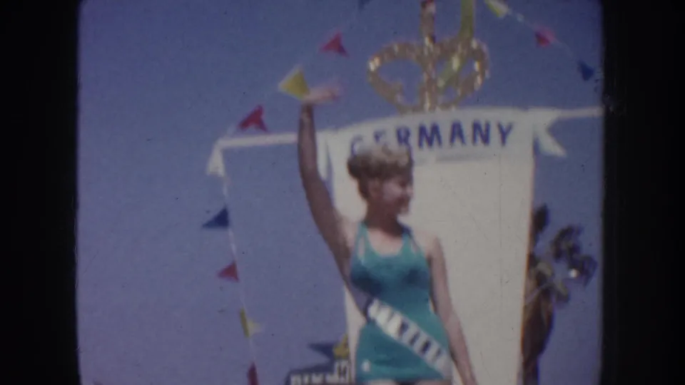 Sash-wearing pageant contestant on a Germany-themed float waves under pennant flags in 1952 California home-movie footage.