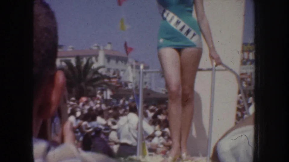 1952 California home-movie frame showing a swimsuit contestant on a parade float, with spectators, railings, and palm trees behind her.