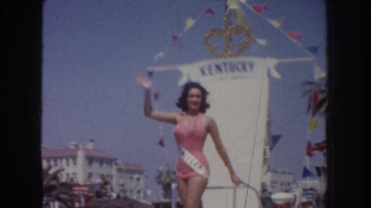 1952 California home-movie frame showing a pageant contestant in a pink swimsuit and sash waving beside a KENTUCKY float sign.