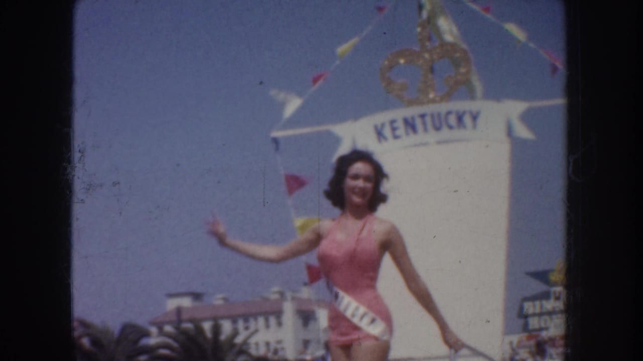 Pink-swimsuit contestant waves in front of a tall KENTUCKY float panel.