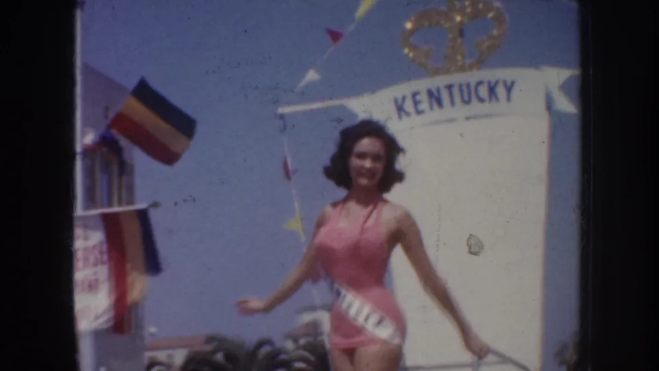 1952 California home-movie frame showing a pink-swimsuit pageant contestant near KENTUCKY signage, flags, and parade decorations.