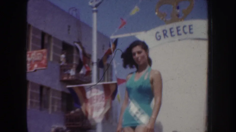 1952 California home-movie frame with a pageant contestant in a teal swimsuit and sash standing beside a GREECE float sign.