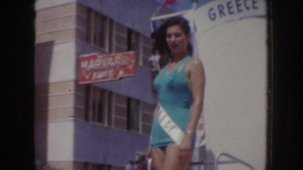 1952 California home-movie frame showing a pageant queen in turquoise with a white sash beside a GREECE-themed float backdrop.