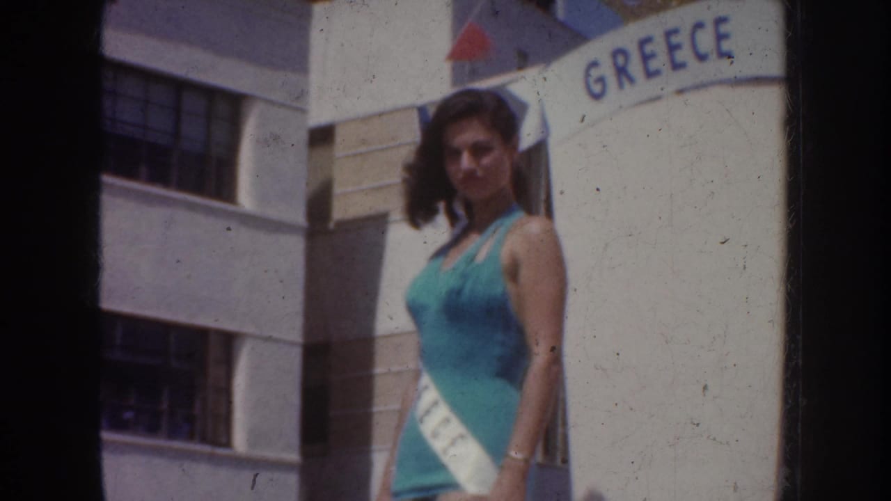 Home-movie frame of the Grecian pageant queen standing in bright sun beside the GREECE lettering and a modern building facade.