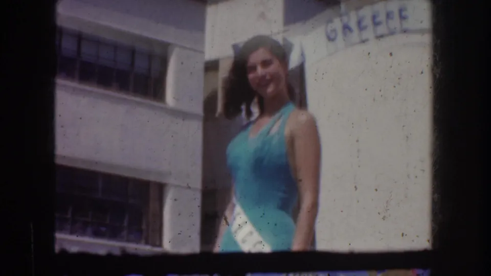 1950s color 8mm frame showing the pageant queen smiling on or near a GREECE-themed parade float in California.