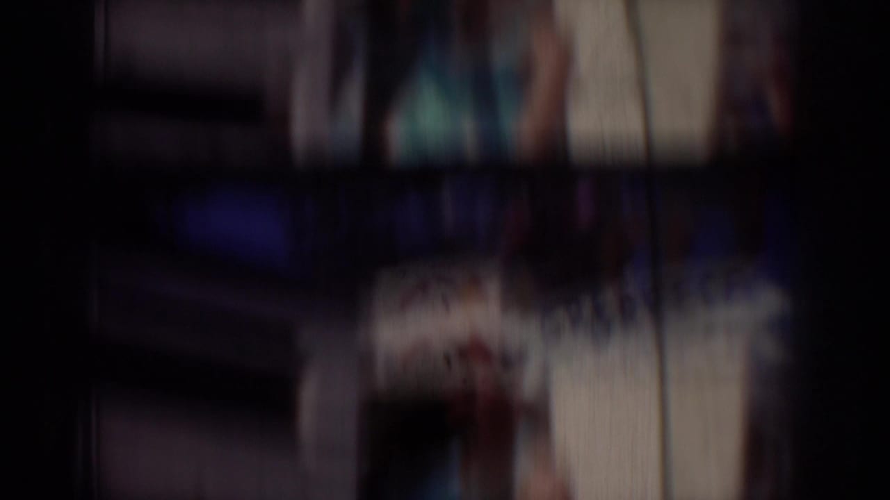 Soft, blurred 1950s 8mm frame from the California parade clip, with dark film edges and indistinct outdoor structures.