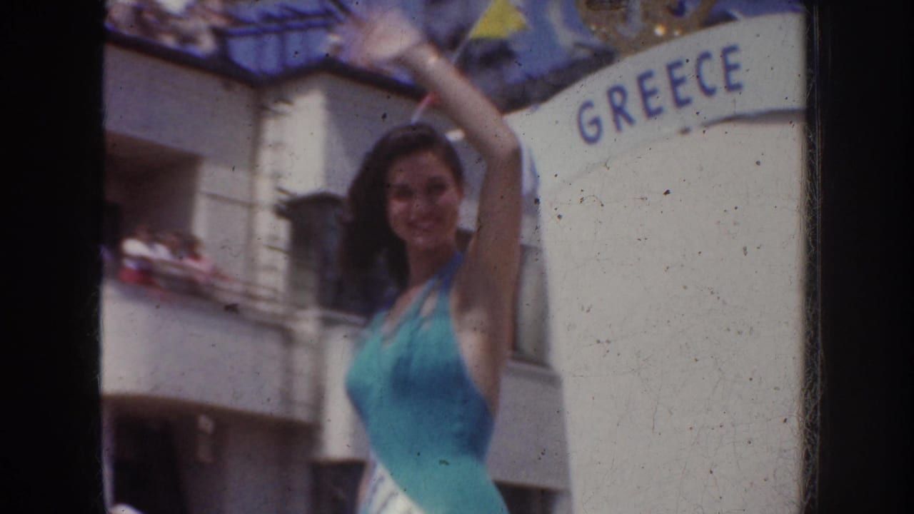 Woman in a turquoise swimsuit waves toward the camera beside a Greece display in the 1952 California parade footage.