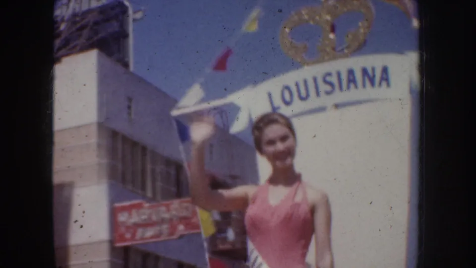 Woman in a pink swimsuit stands and waves near a Louisiana booth in the 1952 California parade footage.