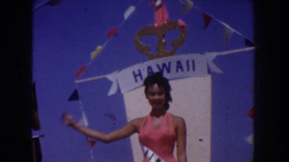 Woman wearing a sash and pink swimsuit waves beside a Hawaii display in bright daylight in the 1952 California footage.