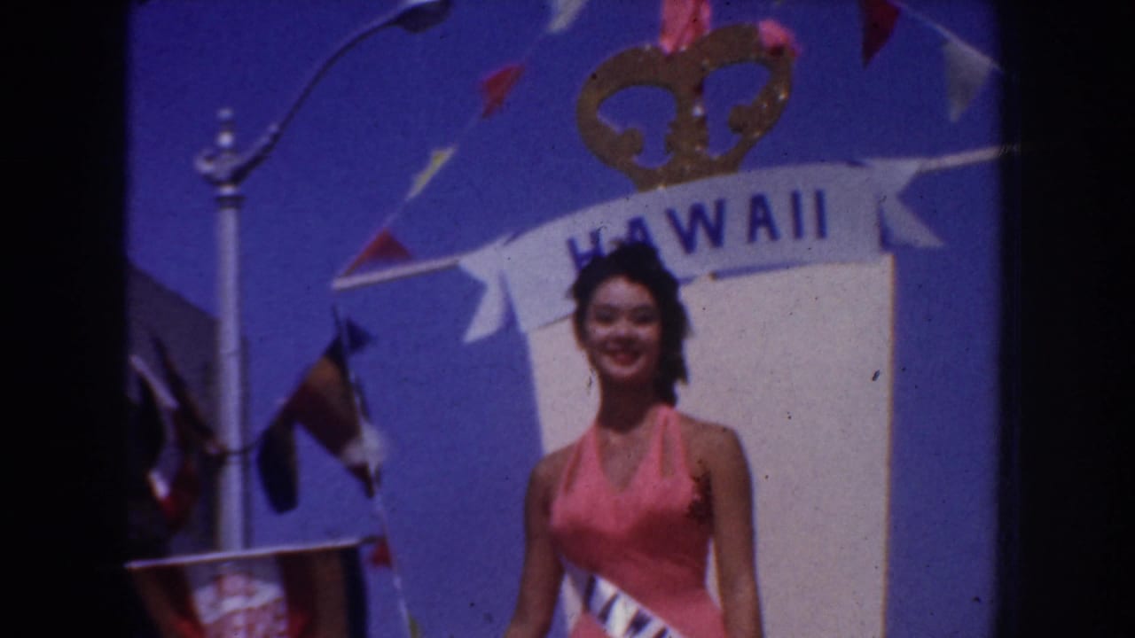 1952 California home-movie frame showing a woman in a pink halter dress and sash beneath a Hawaii banner with flags overhead.