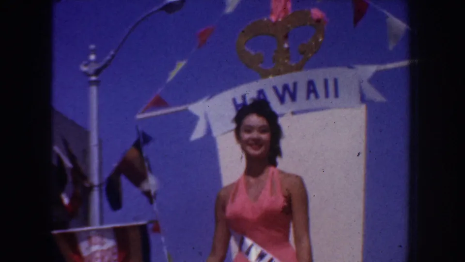 1952 California home-movie frame showing a woman in a pink halter dress and sash beneath a Hawaii banner with flags overhead.