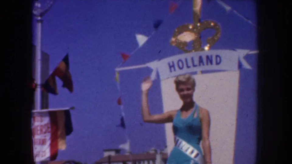 1952 California parade frame showing a blonde woman in a blue dress and sash beneath a Holland banner and crown decoration.