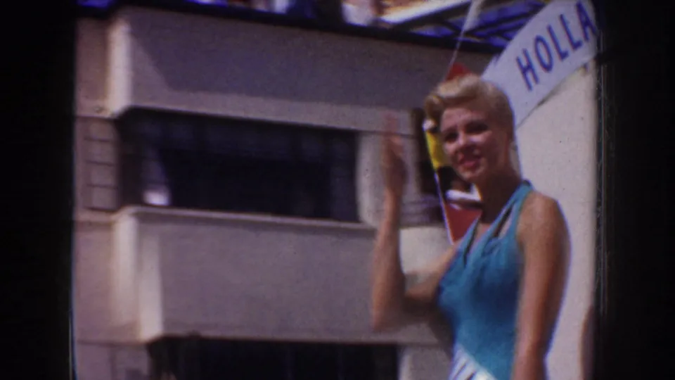 Blonde pageant contestant in a blue dress waves beside a partial Holland banner in 1952 California home-movie parade footage.