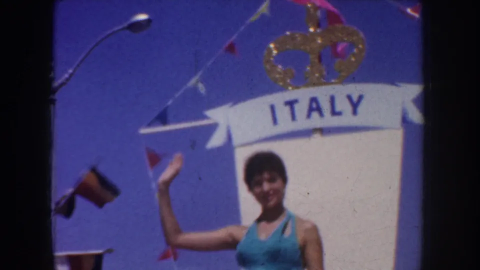 A turquoise-clad participant waves beneath an ITALY banner and gold crown decoration in 1952 California parade footage.