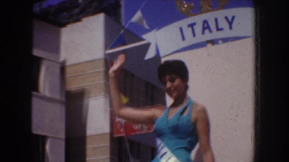 A smiling pageant participant waves next to the ITALY float sign, with bunting and sunlit buildings visible behind her.