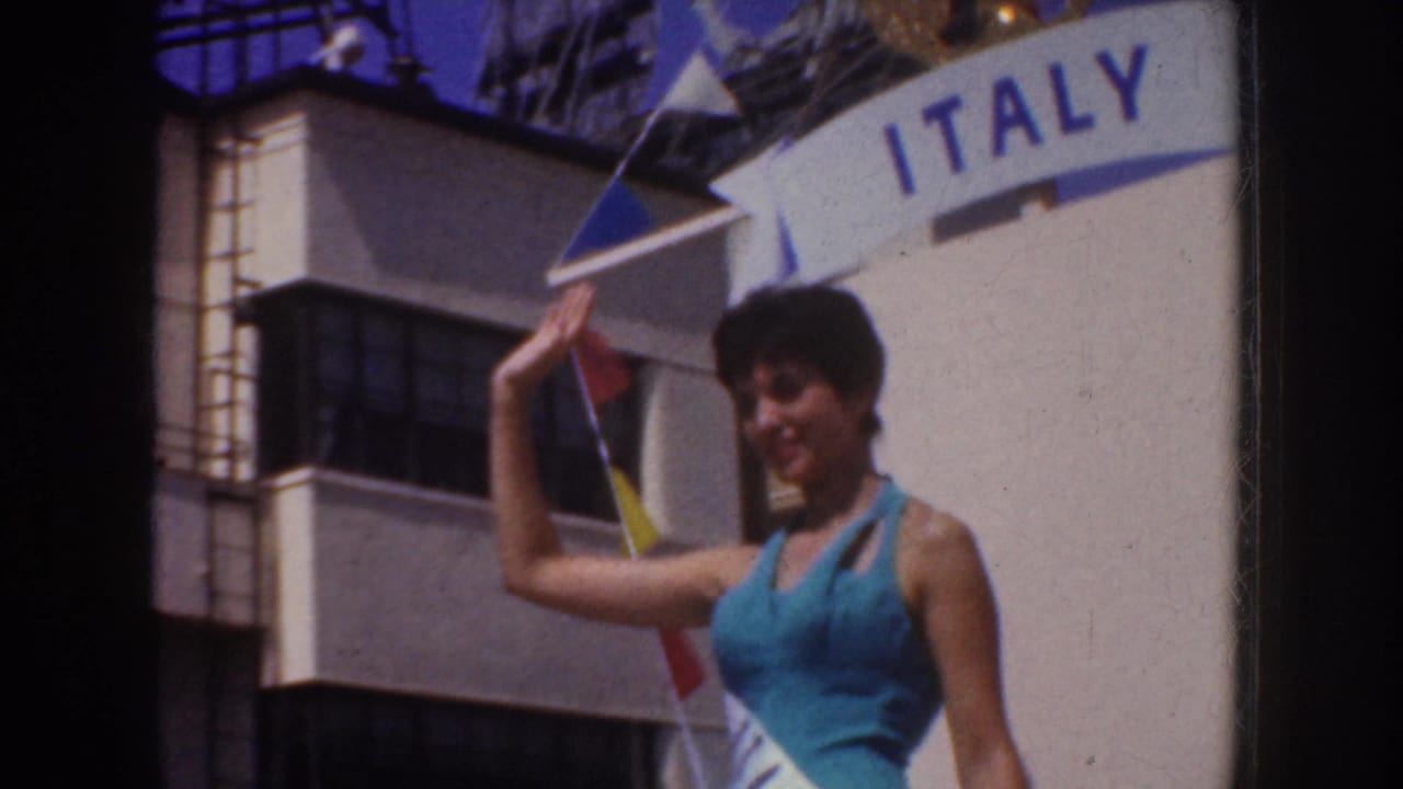 In supplied 1952 California parade footage, a woman in blue waves beside an Italy banner with buildings behind her.