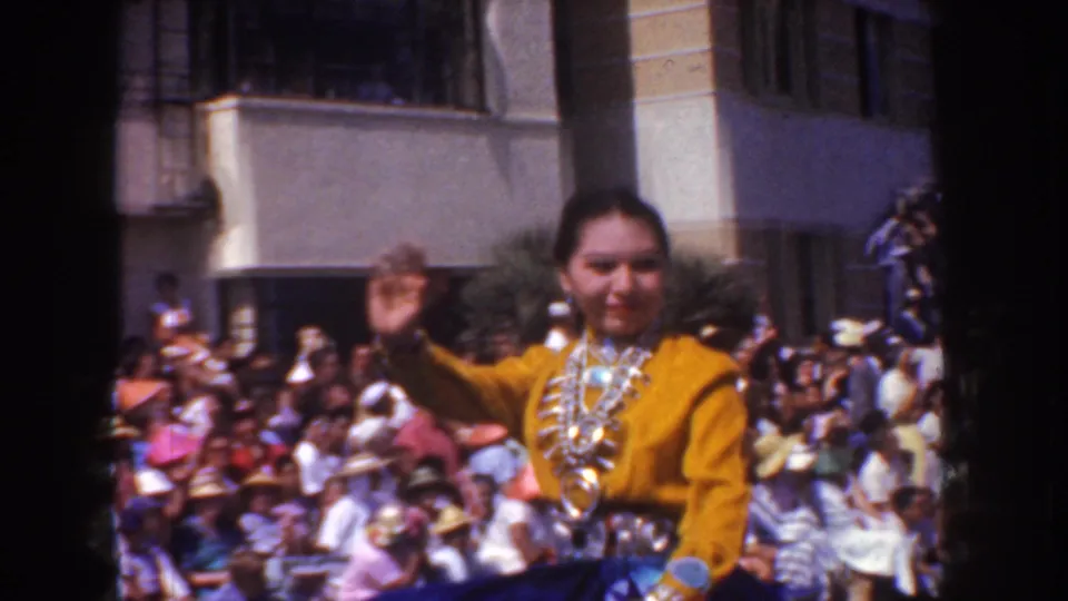 The costumed woman continues waving from the parade route, with a dense seated crowd and building frontage behind her.