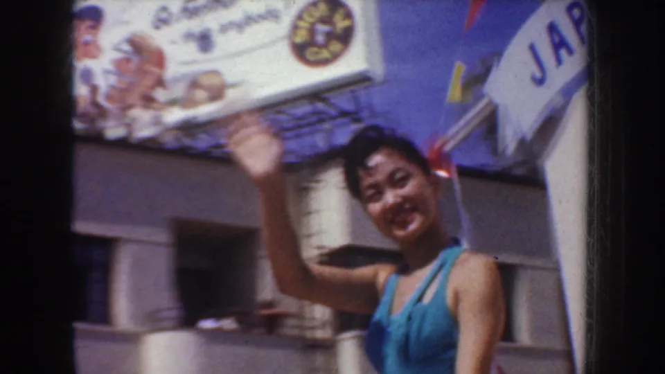 A woman in a blue outfit waves near a Japan banner, with building columns and a partial parade sign visible behind her.