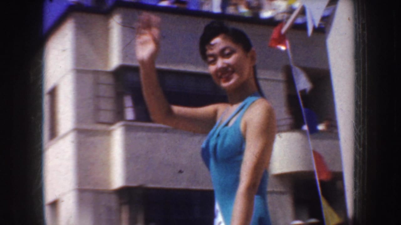 In 1952 California home-movie footage, a smiling woman in a blue swimsuit waves near a sunlit building with small flags overhead.