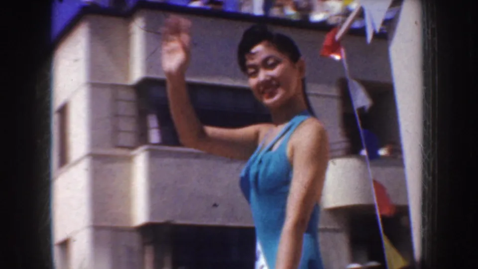 In 1952 California home-movie footage, a smiling woman in a blue swimsuit waves near a sunlit building with small flags overhead.