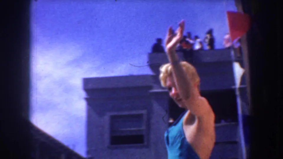 A woman in a blue swimsuit waves in bright sun as spectators stand on a balcony above, seen in handheld 8mm framing.