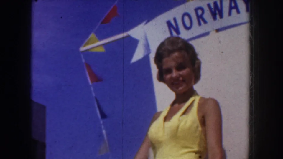 A woman in a yellow swimsuit stands smiling near a large Norway sign, with colorful pennant flags against the sky.