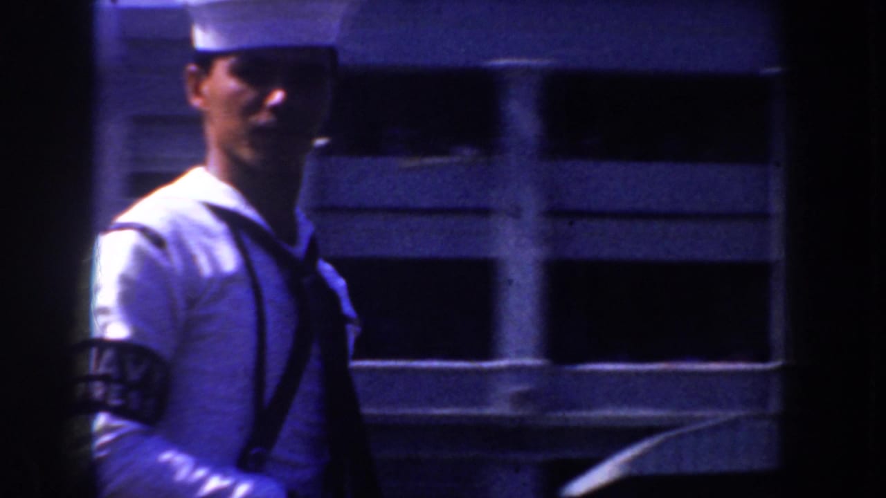 1952 California 8mm frame of a sailor in a white cap and uniform walking near a modern-looking facade with dark window openings.