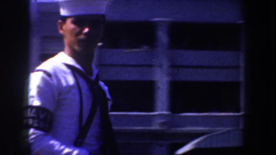 1952 California 8mm frame of a sailor in a white cap and uniform walking near a modern-looking facade with dark window openings.