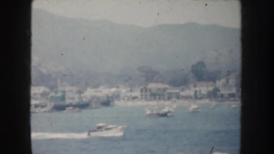 1952 California coastal bay scene with small boats, shoreline buildings, and hills in soft home-movie focus.