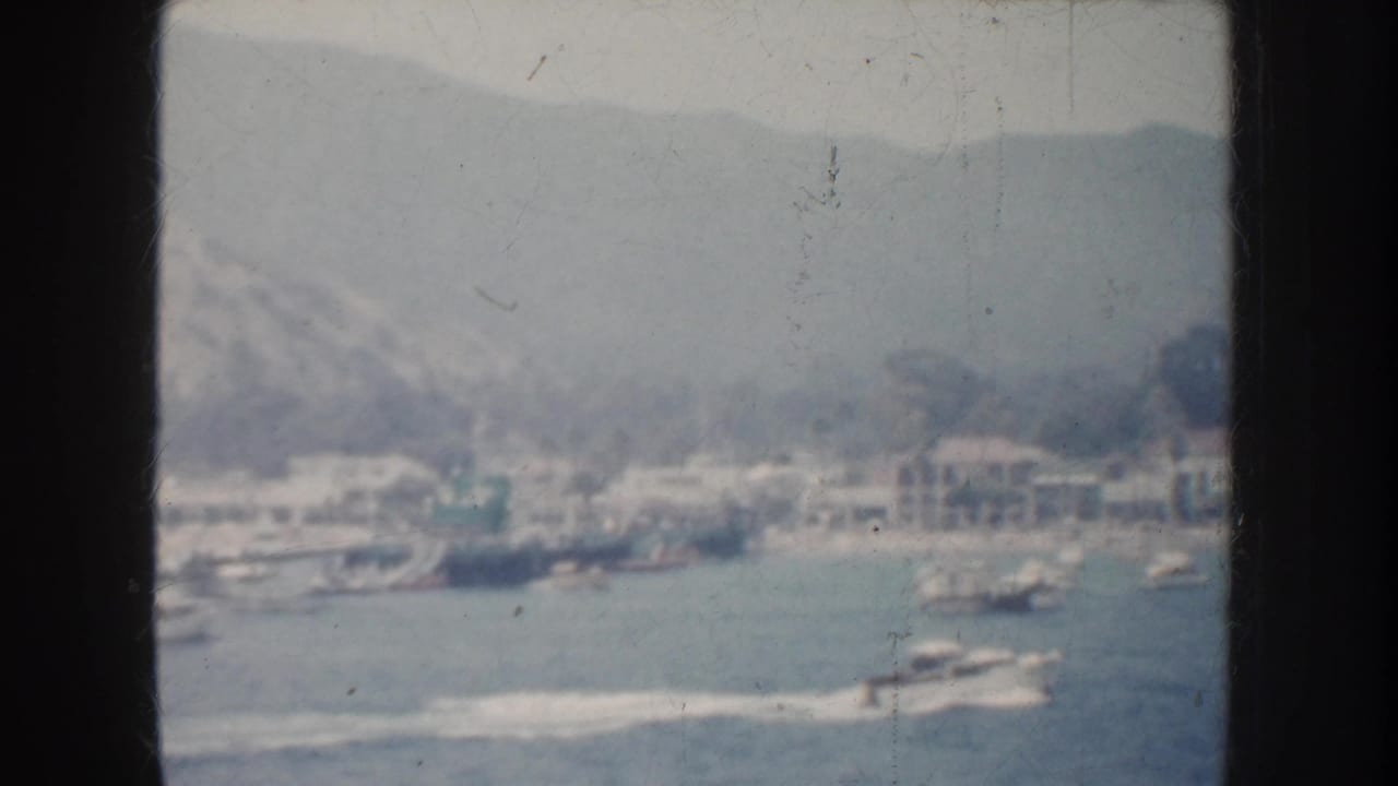 1952 California 8mm view of a coastal harbor town with boats on the water, pale buildings near shore, and hills in the background.