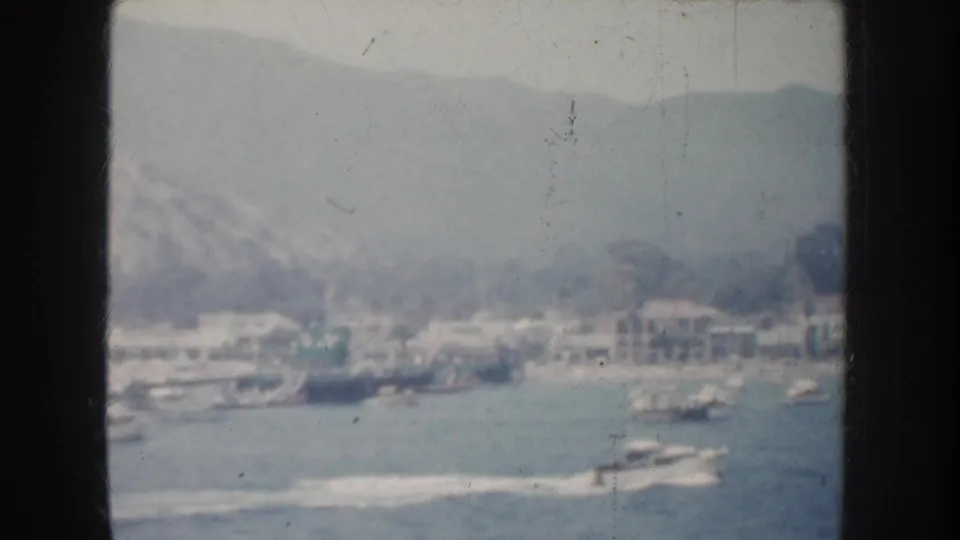 1952 California 8mm view of a coastal harbor town with boats on the water, pale buildings near shore, and hills in the background.