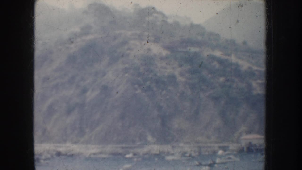 Soft 8mm frame of a California coastal hillside rising over harbor water, with roads or terraces faintly visible.