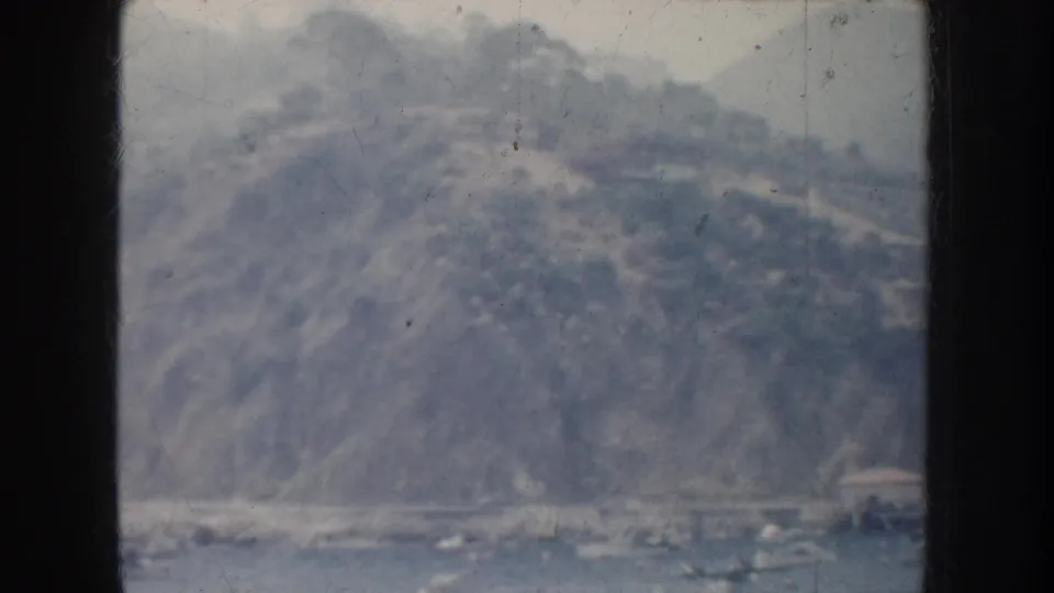 Soft 8mm frame of a California coastal hillside rising over harbor water, with roads or terraces faintly visible.