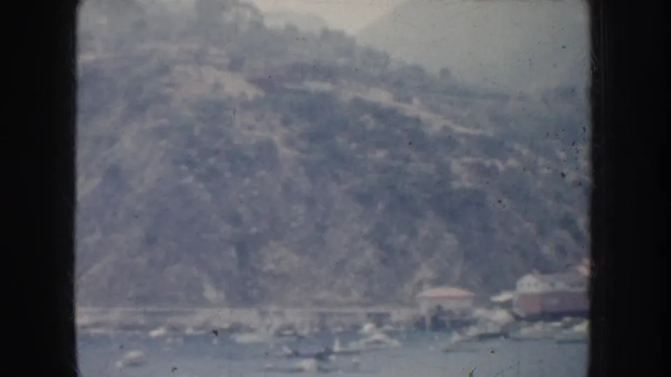 Faded 8mm view of a California harbor shoreline with boats below a steep hillside and small waterfront buildings.