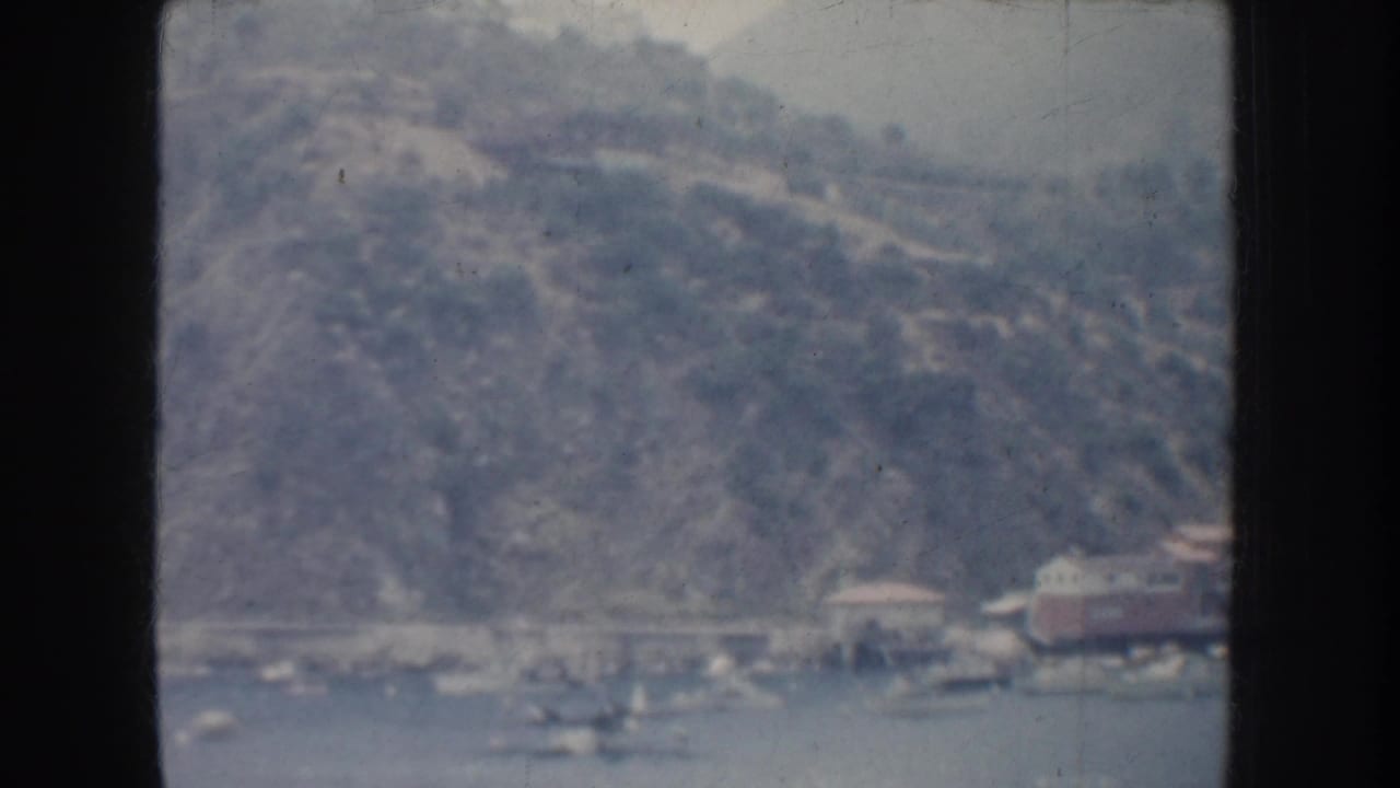 1952 California 8mm frame showing a distant lakeside shore, rugged hillside, small waterfront structures, and boats on the water.