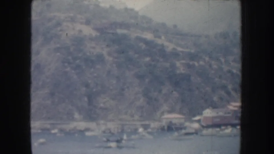 1952 California 8mm frame showing a distant lakeside shore, rugged hillside, small waterfront structures, and boats on the water.