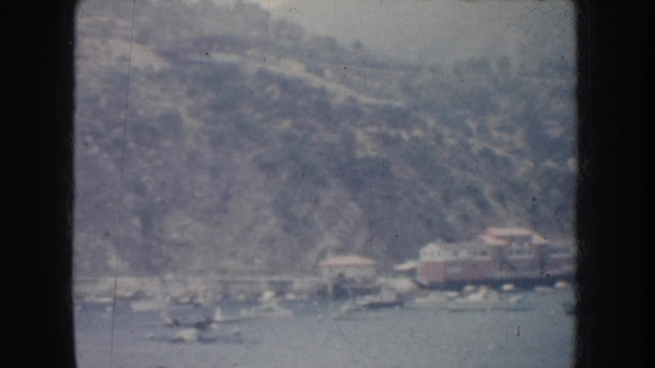 1952 California home-movie frame with a hazy hillside shore, small lakeside structures, and several boats visible on the water.