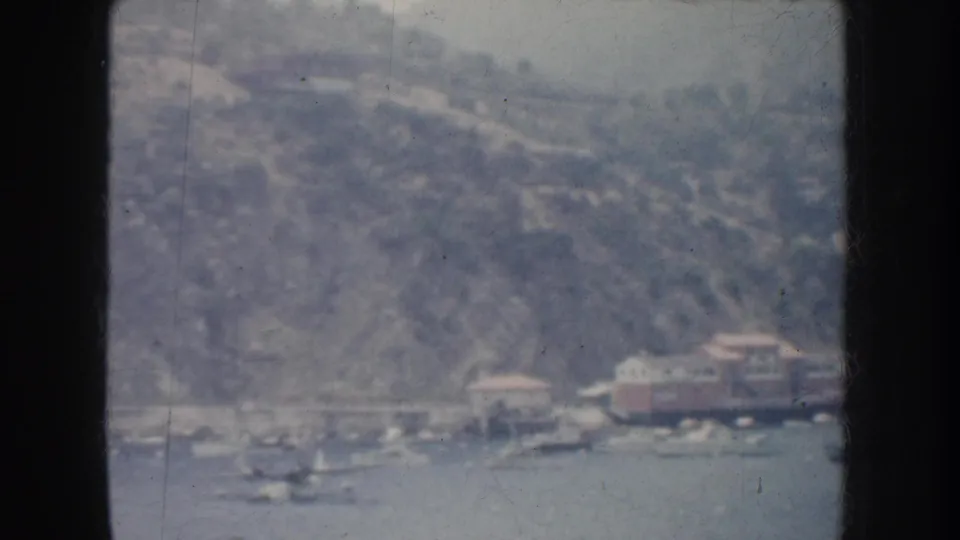 1952 California home-movie frame with a hazy hillside shore, small lakeside structures, and several boats visible on the water.