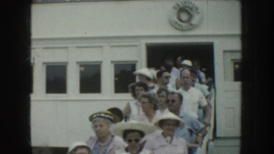 1952 California 8mm frame showing people in summer hats and light clothing exiting a white doorway with an emblem above it.