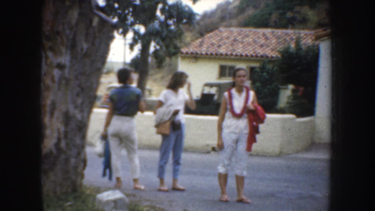 1952 California home-movie frame showing three women visitors near a tree and stucco church exterior, with a tiled roof in the background.