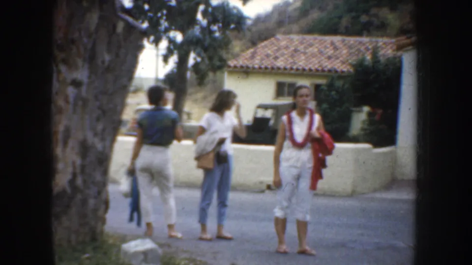1952 California home-movie frame showing three women visitors near a tree and stucco church exterior, with a tiled roof in the background.