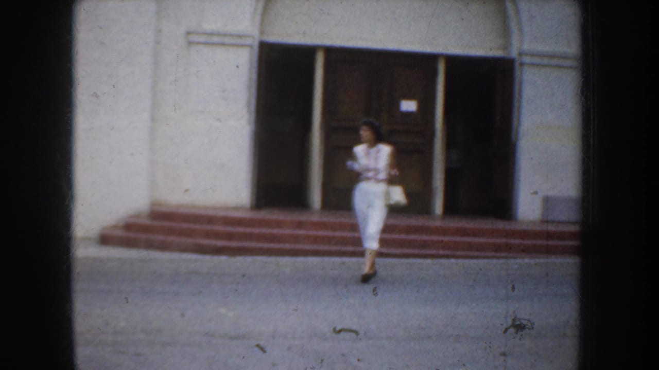 Woman in white walks near the steps of a California church entrance, framed by white stucco walls and dark wooden doors.
