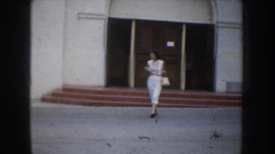 Woman in white walks near the steps of a California church entrance, framed by white stucco walls and dark wooden doors.