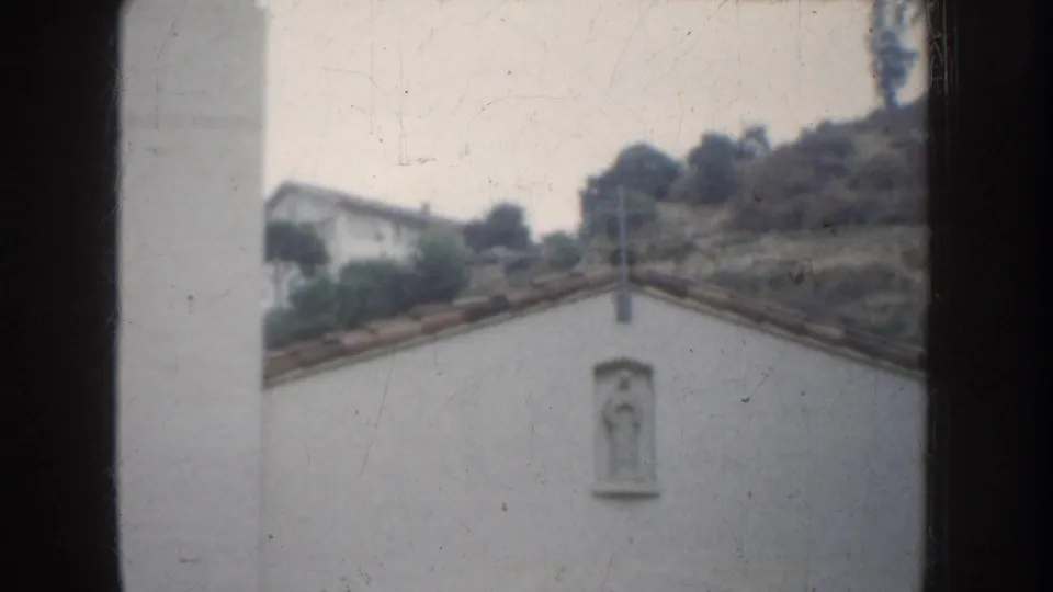 California church exterior detail with a white wall, small cross, statue niche, sloped roofline, and hillside background.