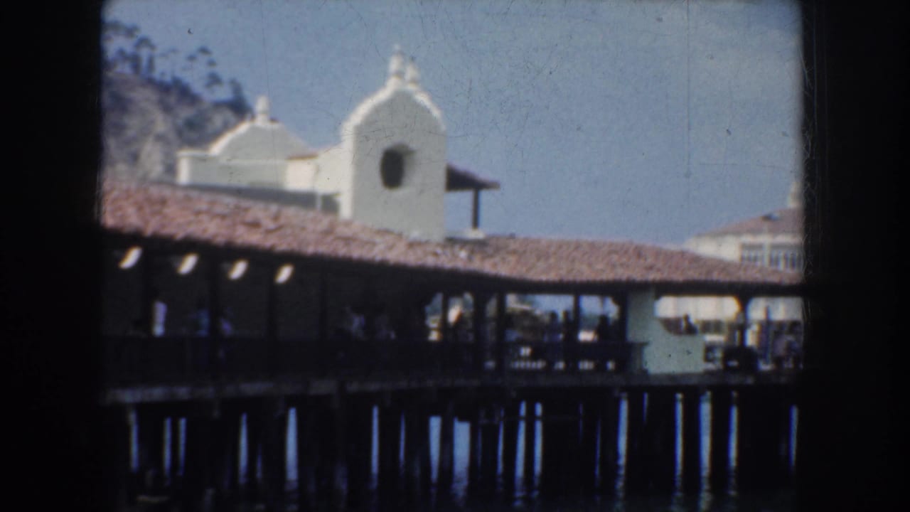 Covered pier walkway over water with a white mission-style tower behind it.