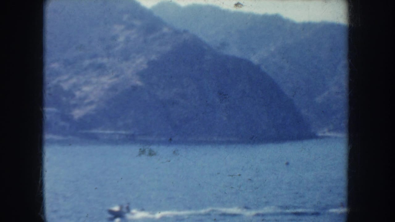 A distant boat crosses water below hazy coastal mountains.