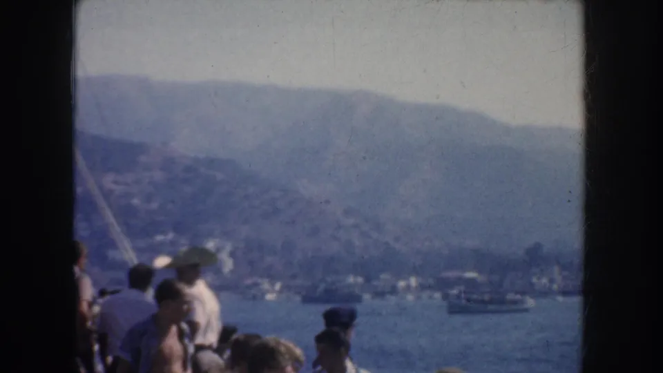 1952 California home-movie frame showing boat passengers in the foreground, calm water, small distant boats, and a hazy mountain backdrop.